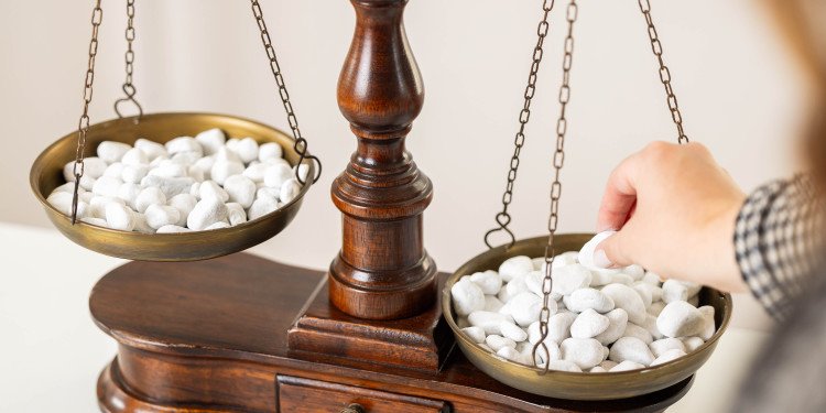 The image shows a balance scale with numerous small, white stones on each pan. A hand is adding another stone.© University of Münster - Roland Baege