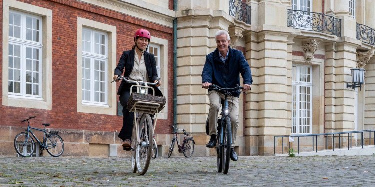 Das Bild zeigt Rektorin Prof. Dr. Susanne Menzel-Riedl und Pressesprecher Norbert Robers, die gemeinsam auf ihren Rädern vom Schloss wegfahren. Sie fahren direkt auf den Fotografen zu. Im Hintergrund ist die Hauptfassade des Schlosses zu sehen.© Uni