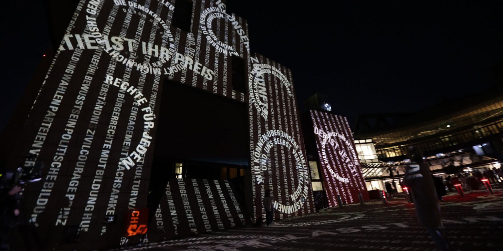 Weiße Schriftzüge mit Zitaten, darunter Worte von Schülern aus Selm, bedecken die Fassade des NRW-Landtags bei Nacht. Die großflächige Lichtprojektion zeigt unterschiedlich große Buchstaben und Wörter, während im Vordergrund einzelne Menschen stehen.