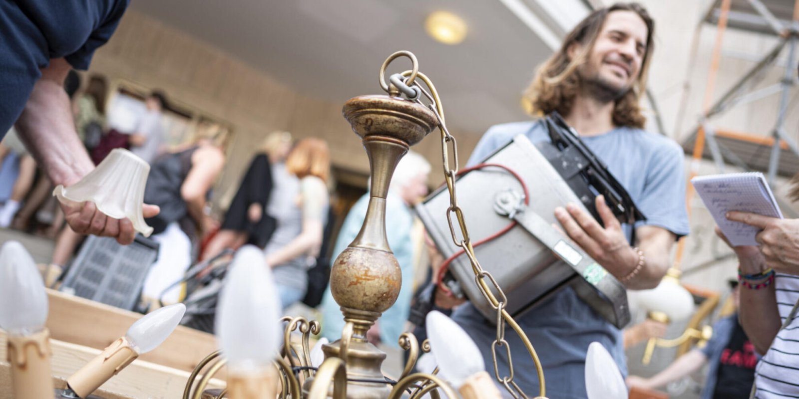 Trödelmarkt mit einem Kronleuchter im Vordergrund und mehreren Personen im Hintergrund.